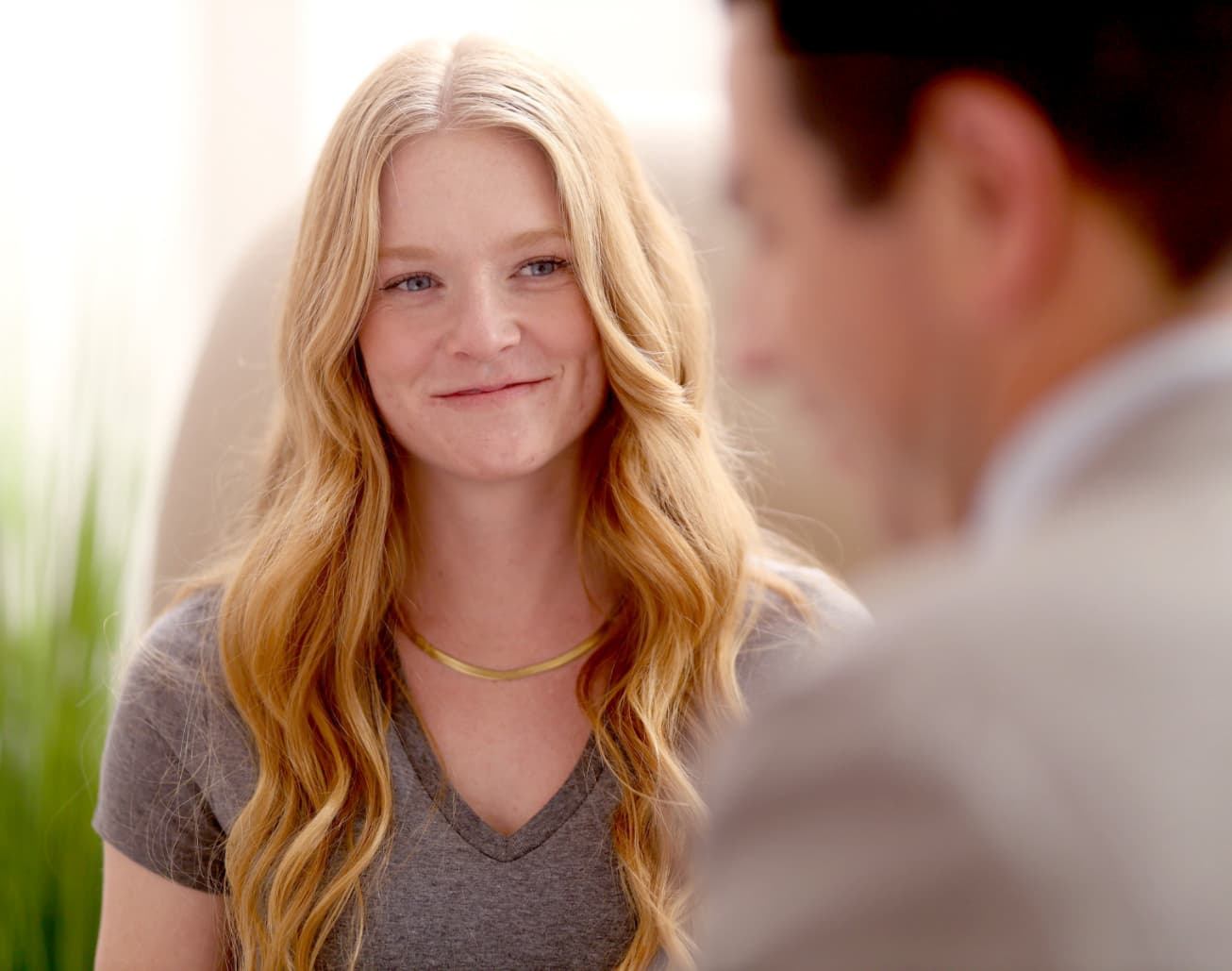Smiling woman with long wavy blonde hair wearing a gray V-neck shirt, sitting indoors and looking at a man in the foreground.