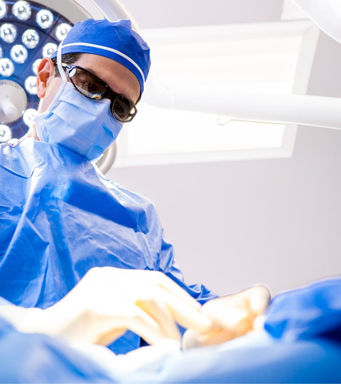 Surgeon wearing a blue surgical gown, mask, cap, and protective glasses performing a procedure under operating room lights.