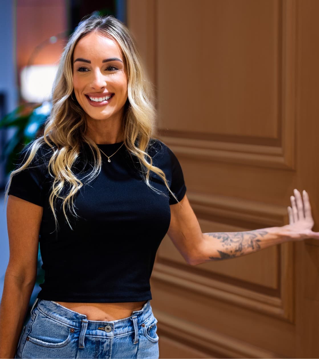 Woman with long blonde hair wearing a black crop top and blue jeans, smiling while standing in a doorway with one hand on the wooden door.