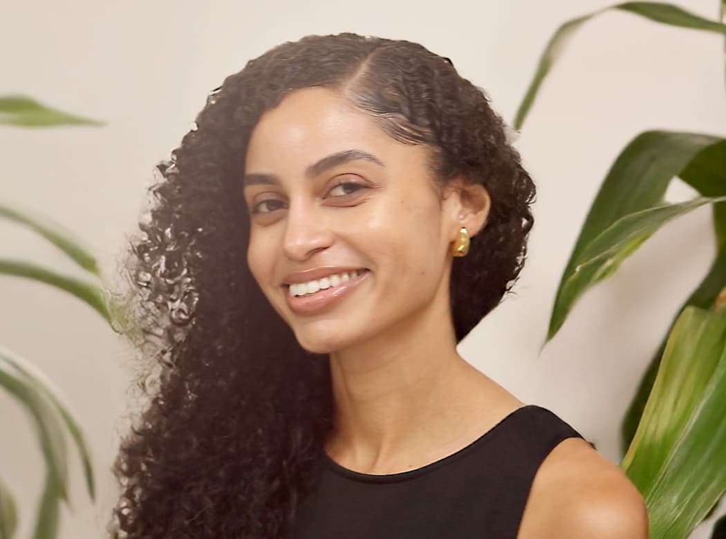 Woman with long curly hair wearing a sleeveless black top, smiling while seated in front of leafy green plants indoors.