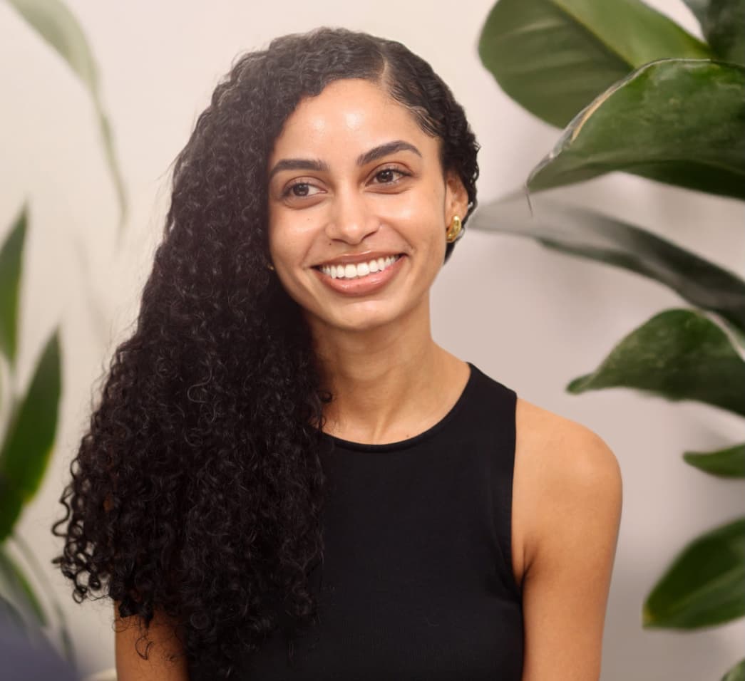 Woman with long curly hair wearing a sleeveless black top, smiling while seated in front of leafy green plants indoors.