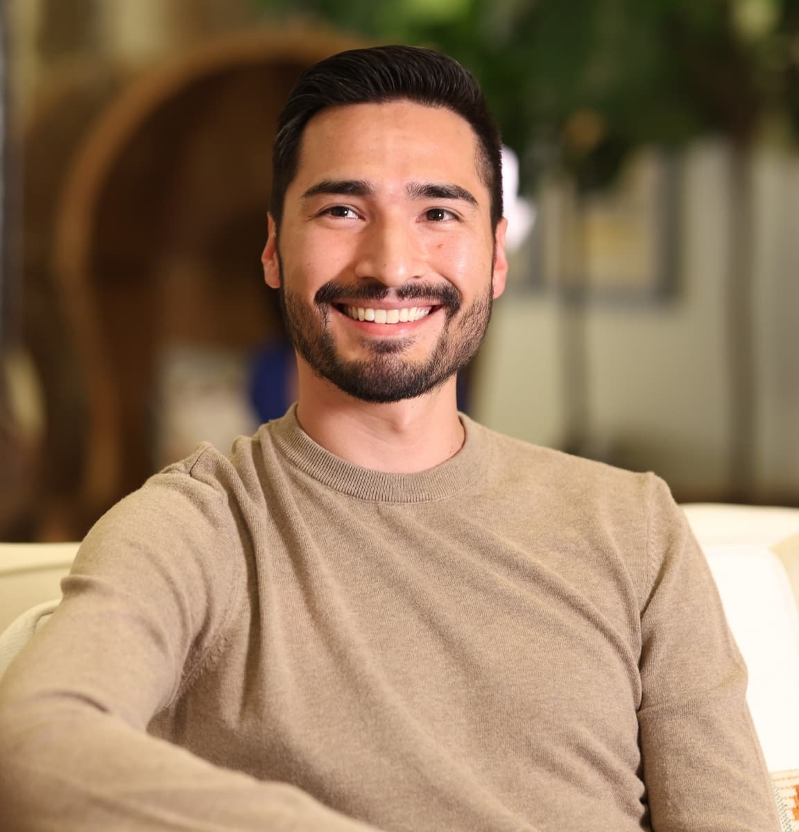 Man with short dark hair and trimmed beard wearing a tan sweater, smiling while seated on a light-colored sofa with a blurred indoor background and greenery.