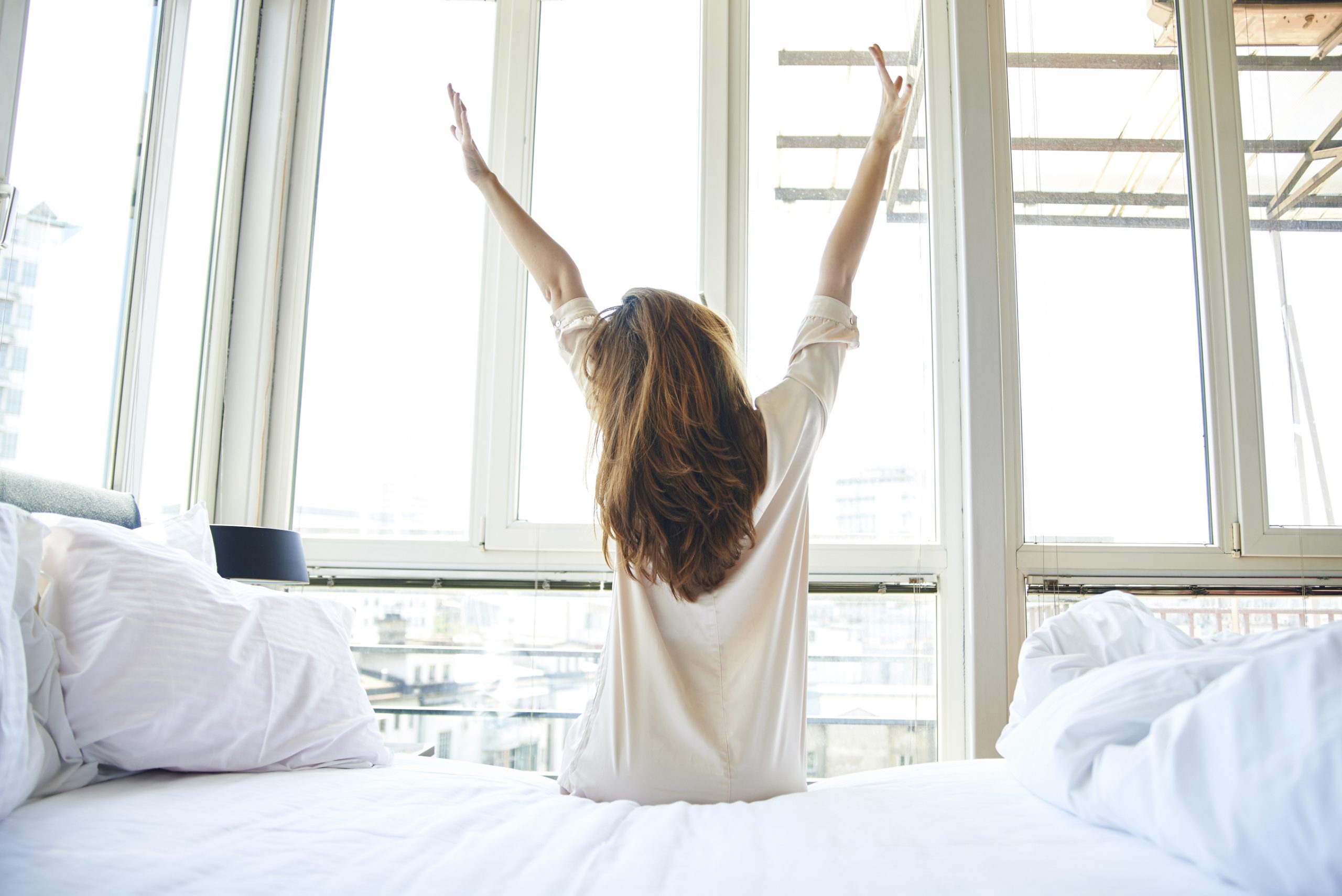 Young woman is doing morning stretching in bed, arms raised, rear view