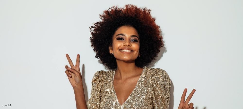 African American woman wearing gold sequined dress and smiling showing peace signs with both hands