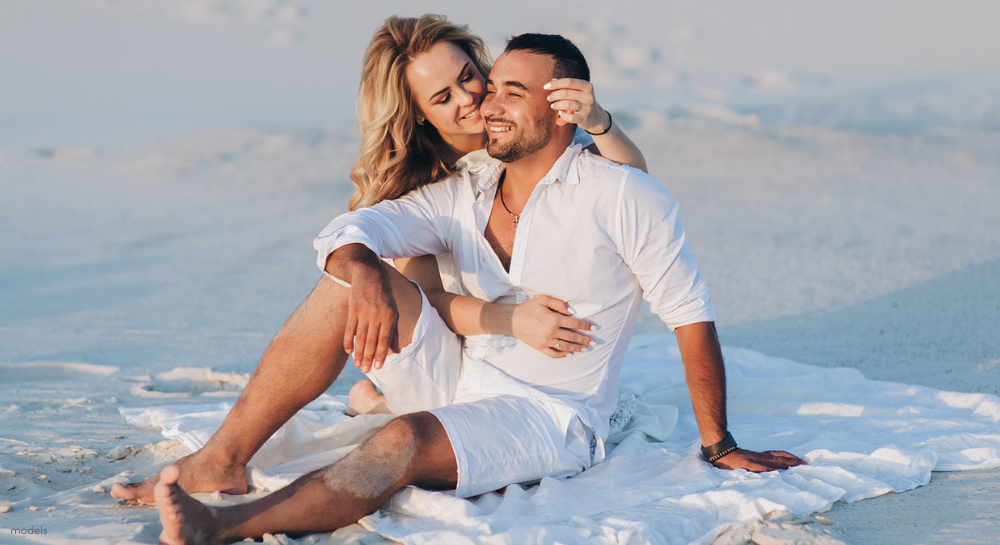 Couple dressed in white sitting on the beach cuddling and enjoying the sunset