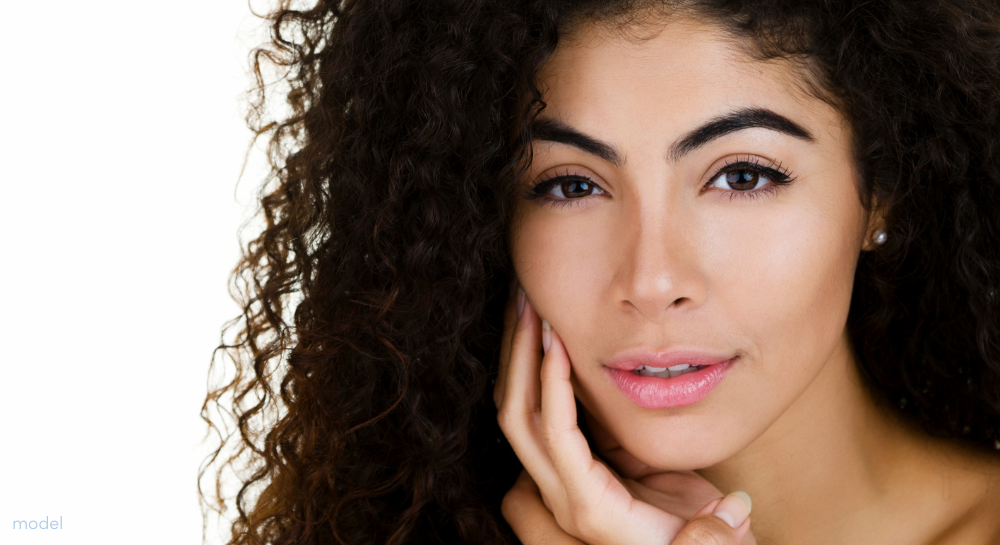 Beautiful curly-haired brunette on white background staring at camera - hand on her face.