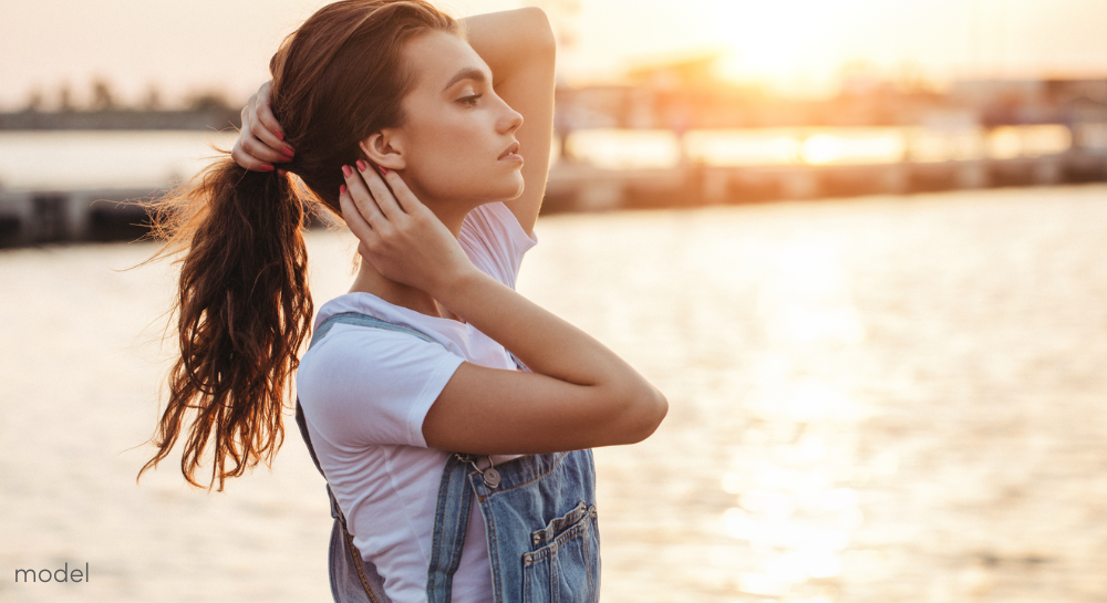 Woman at the docks on the water, holding her hair in a pony tail and touching her neck