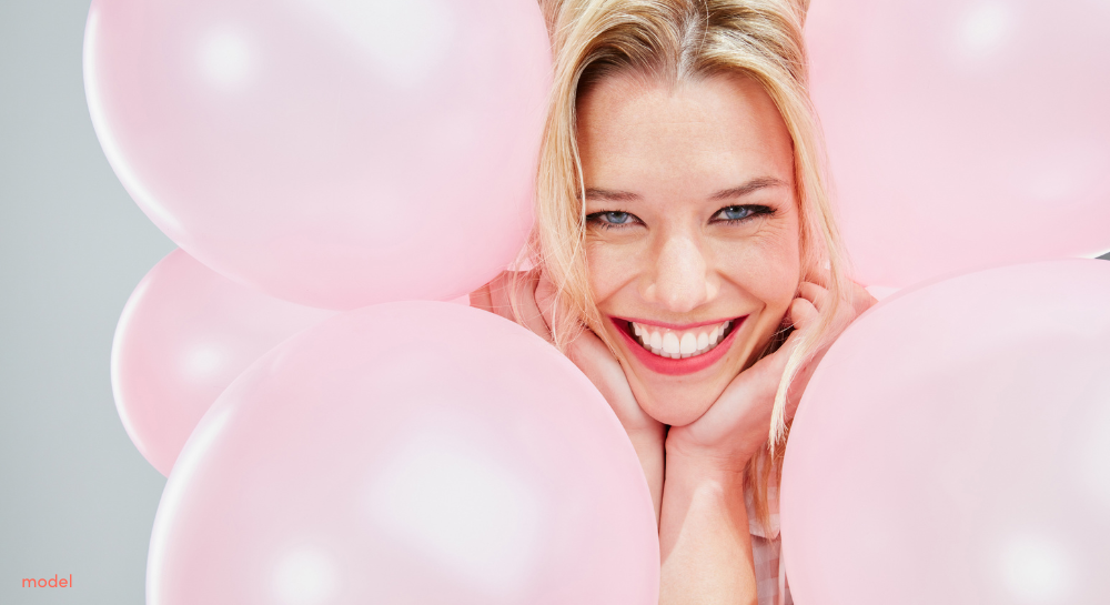 Closeup of blonde woman smiling at the camera, hands on her chin, surrounded by pink balloons