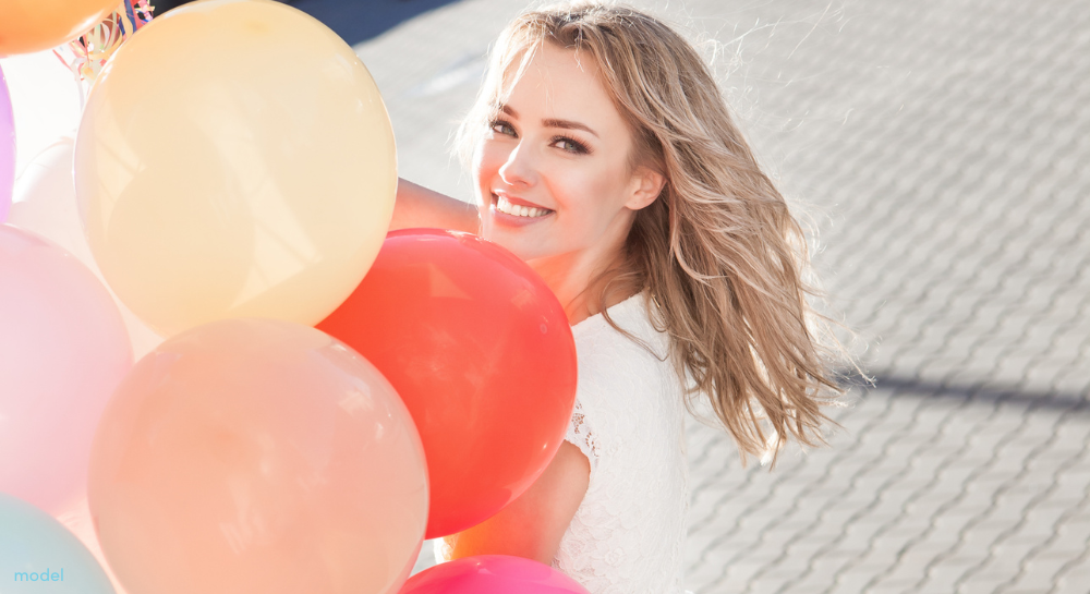 Pretty blonde walking outdoors holding a bunch of colorful balloons and smiling.