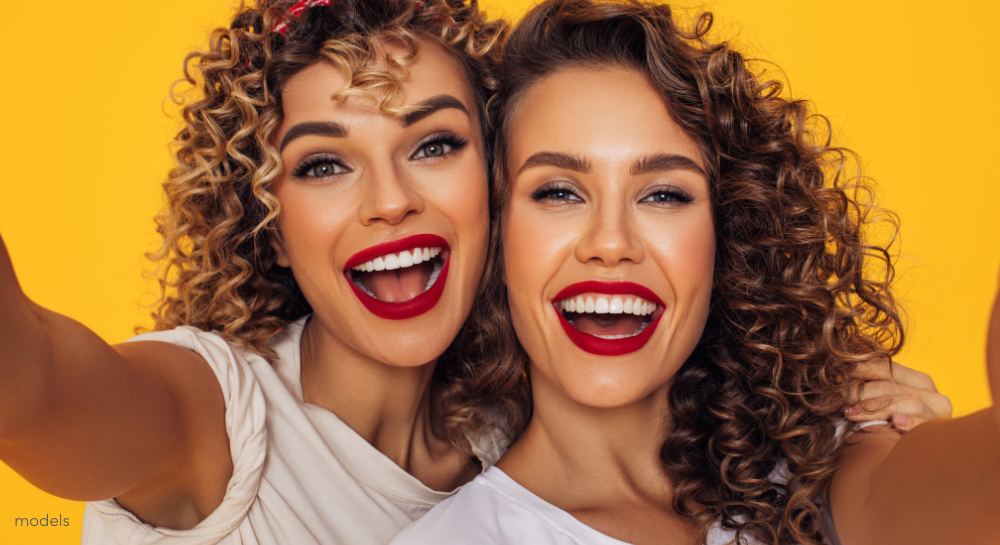 Two women with red lipstick and curly hair taking a selfie against a golden yellow background