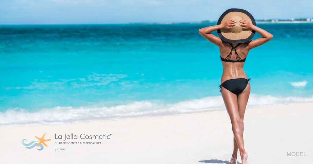 A fit woman standing on the beach in a black bikini. Her arms are up and holding on to her wide-brim beach hat. (Model)