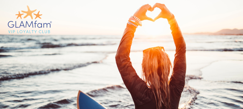 Female surfer on a board holding her hands into qa heart shape above her head, looking out to the sunset.
