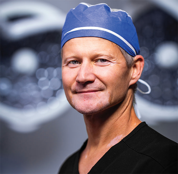 Dr. Lukasz “Luke” Swistun, board-certified plastic surgeon at LJCSC – La Jolla Cosmetic Surgery Centre – smiling confidently in surgical attire under bright OR lights.