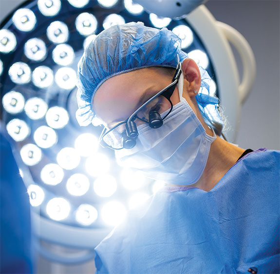 Board-certified plastic surgeon at LJCSC – La Jolla Cosmetic Surgery Centre – performing surgery with precision under bright operating lights, wearing surgical loupes, mask, and sterile.