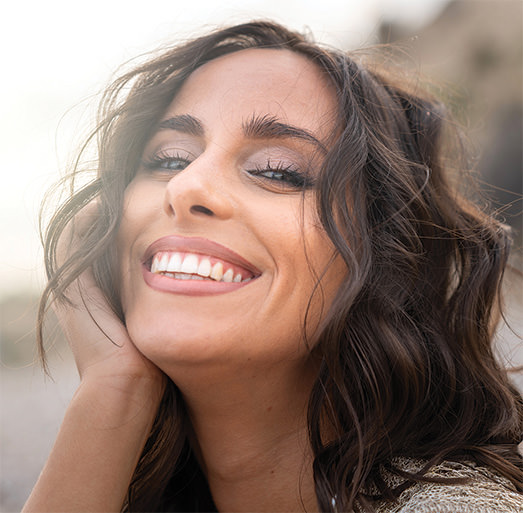 Woman with wavy dark brown hair smiling with her head tilted and hand resting on her cheek, photographed outdoors with a blurred background.