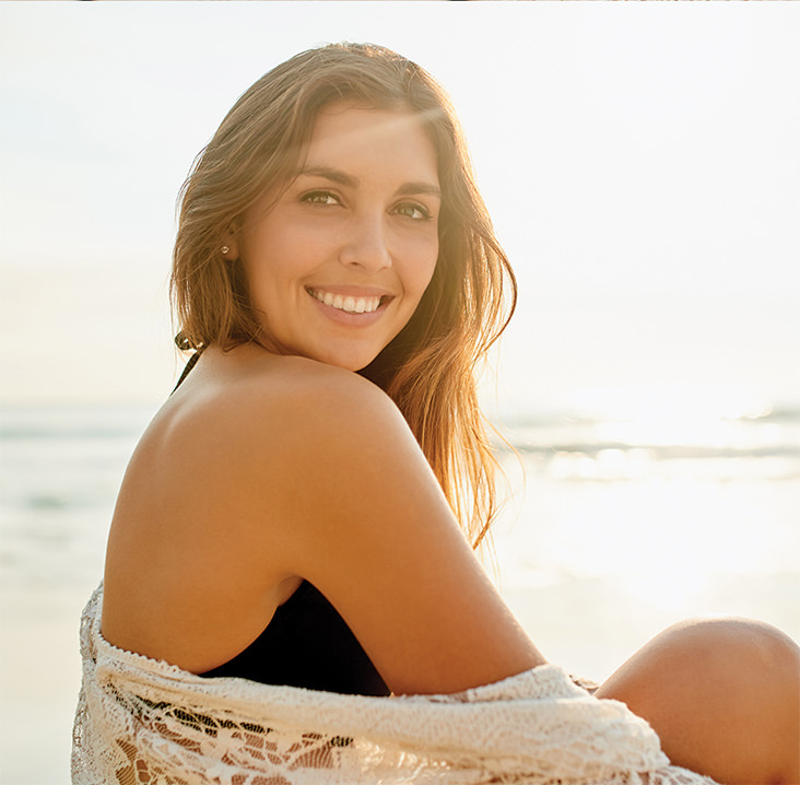 Woman with long brown hair sitting on the beach at sunset, wearing a black swimsuit and lace cover-up, smiling over her shoulder with the ocean in the background.