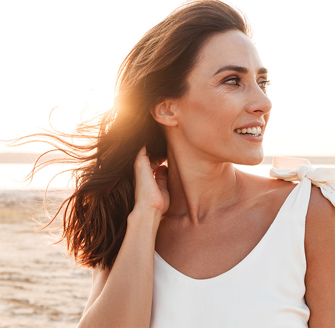 Woman with long brown hair wearing a white sleeveless top, standing on the beach at sunset with her hand touching her hair and wind blowing through it.