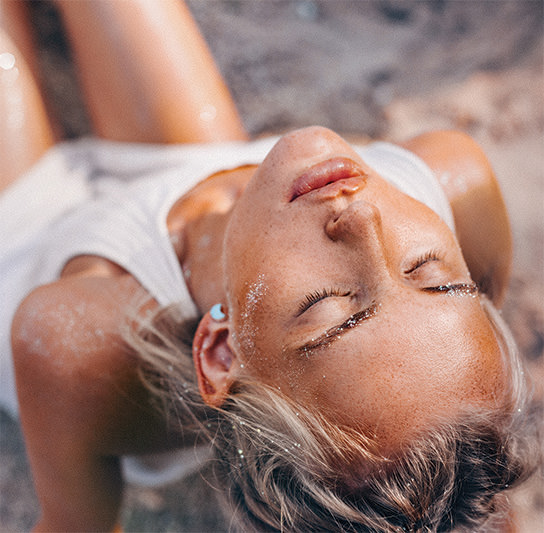 Woman with sandy skin and glitter on her eyebrows reclining on the beach with eyes closed, wearing a white tank top and light blue ear stud.