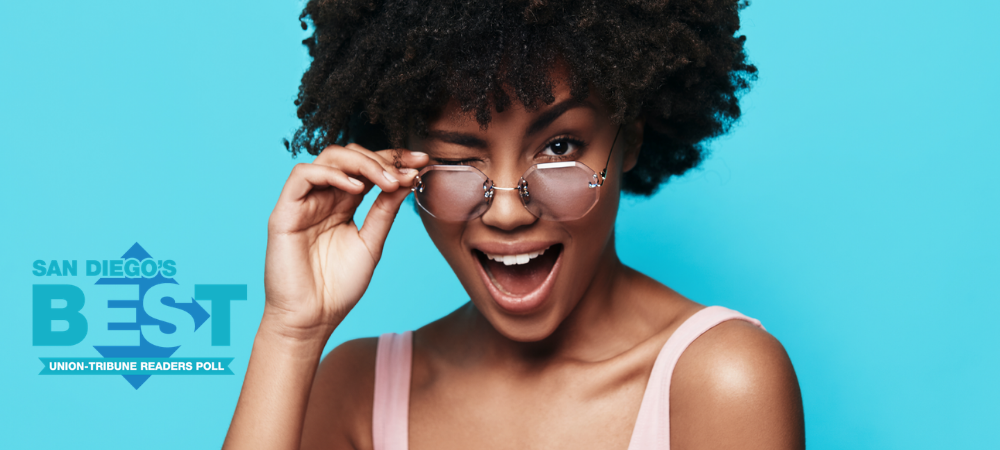 Smiling woman with natural curly hair wearing a light pink tank top and tinted glasses, playfully winking while adjusting her glasses against a bright blue background, with the San Diego’s Best Union-Tribune Readers Poll logo on the left.