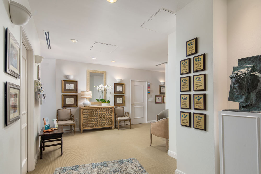 Interior view of La Jolla Cosmetic Surgery Centre reception area with beige carpet, framed certificates on the walls, a console table with white orchids and a lamp, cushioned chairs, and a wall displaying multiple award plaques.