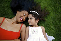 Smiling woman in a red top lying on grass next to a young girl in a white dress, looking at each other affectionately.