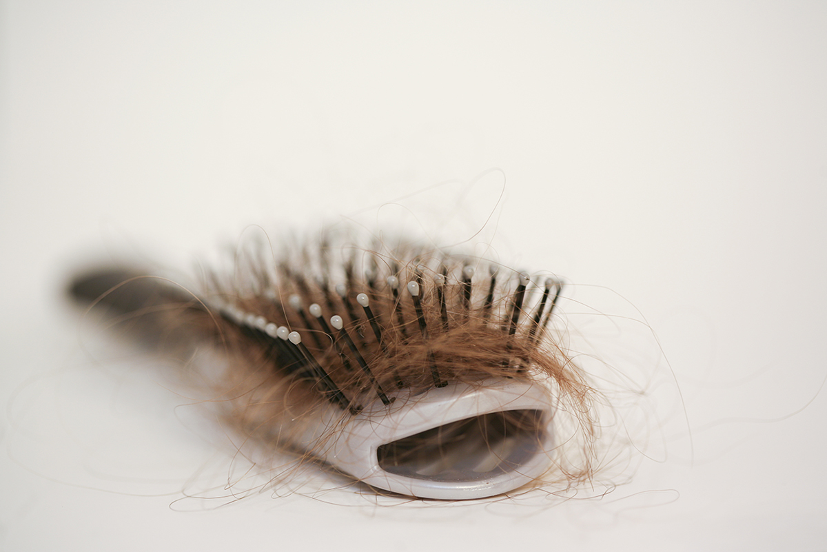 Close-up of a hairbrush with strands of light brown hair tangled in the bristles, set against a plain background.