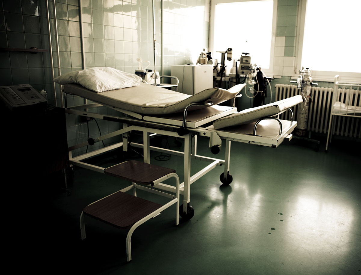 Empty hospital examination room with a padded medical bed on wheels, a pillow, step stools, medical equipment, oxygen tanks, and tiled walls, lit by natural light from a window.