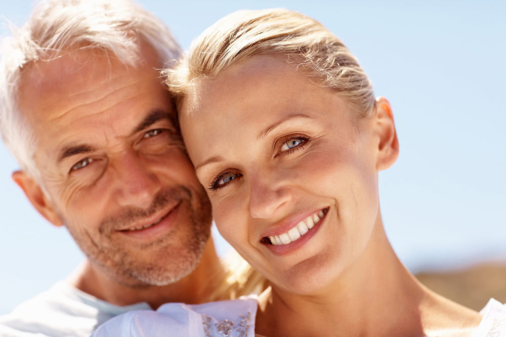 lip lift san diego Close-up of a smiling middle-aged couple with the man resting his head against the woman’s, both looking at the camera with a clear blue sky in the background.
