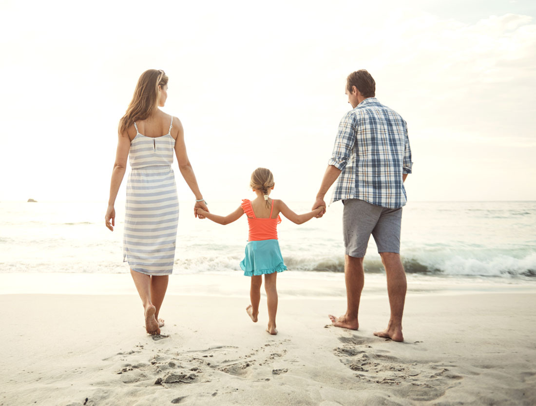 vaginal rejuvenation Man and woman walking barefoot on the beach holding hands with a young girl between them, facing the ocean waves under a bright sky.