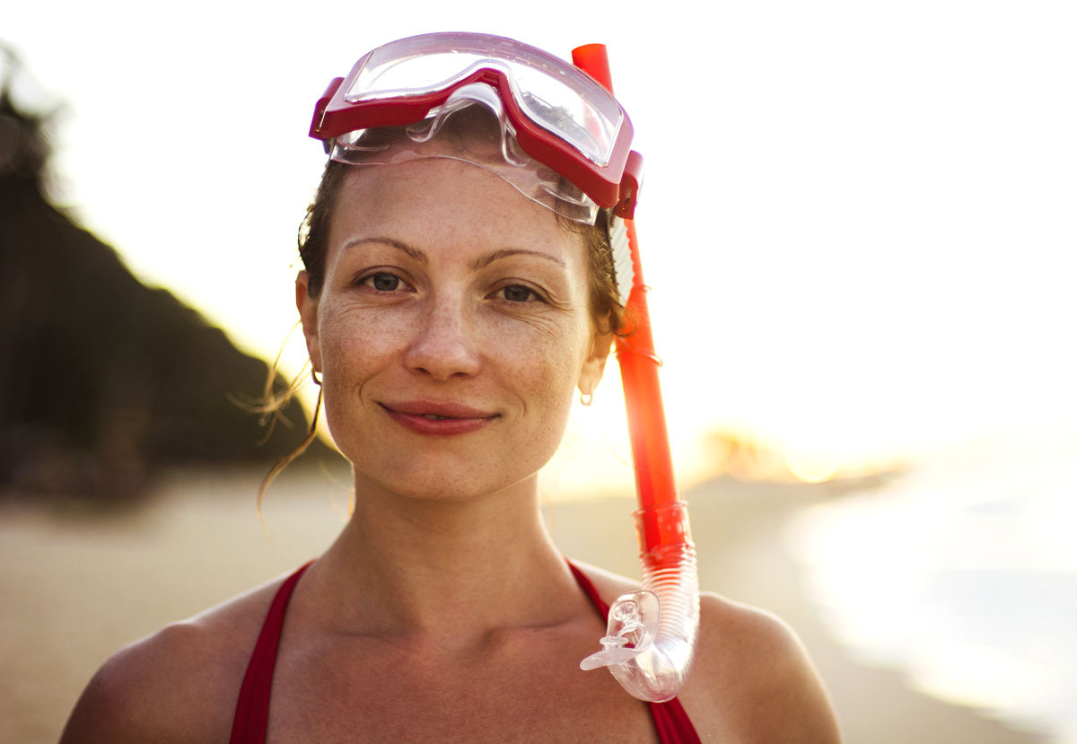 Woman wearing a red snorkel mask and snorkel, standing on a beach at sunset with the ocean and shoreline in the background.