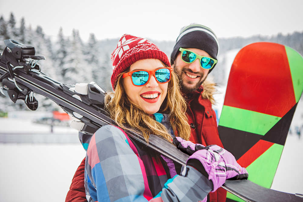 Smiling woman in red winter hat and colorful ski jacket carrying skis on her shoulder, standing next to a man in a black beanie holding a snowboard with red, green, and black stripes, both wearing reflective sunglasses on a snowy mountain.