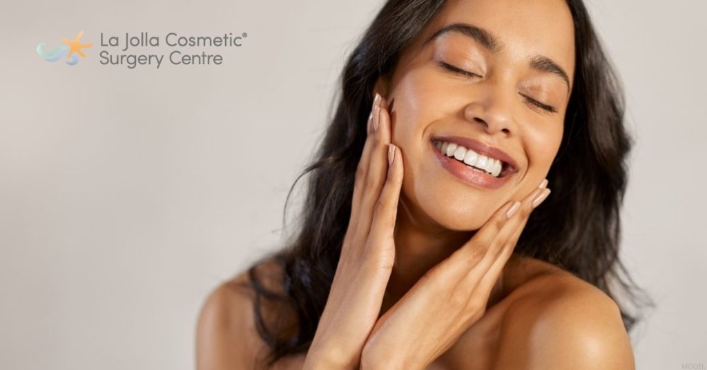 Young woman is smiling and holding her hands to her face (model) La Jolla Cosmetic Surgery Centre Logo